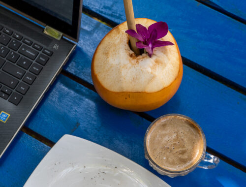 blue table with a cake, king coconut, wooden straw and orchid flower, coffee with foam and laptop open